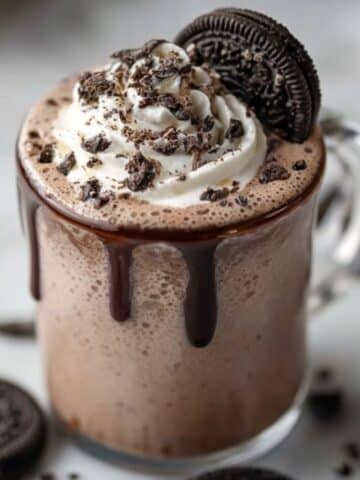 Oreo hot chocolate in a clear glass mug with chocolate drizzle, topped with whipped cream, crushed Oreo cookies, and a whole Oreo on top.