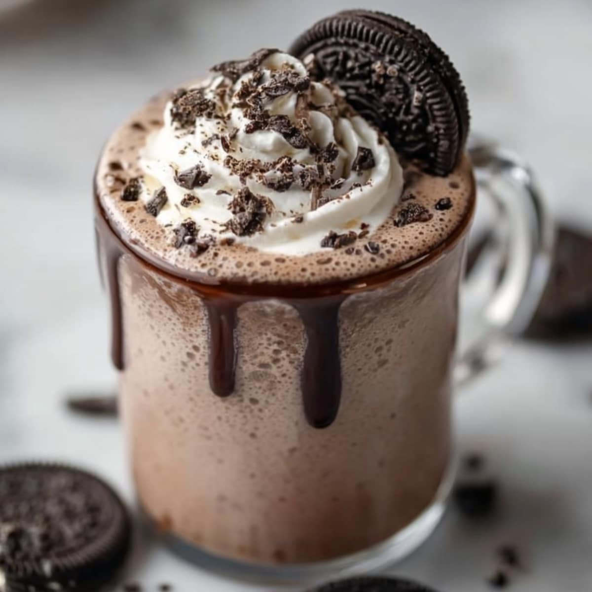 Oreo hot chocolate in a clear glass mug with chocolate drizzle, topped with whipped cream, crushed Oreo cookies, and a whole Oreo on top.