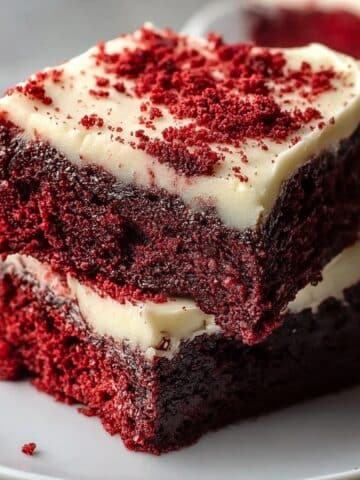 Red velvet brownies with thick cream cheese frosting, rich fudgy layers, and red velvet crumbs sprinkled on top, served on a white plate