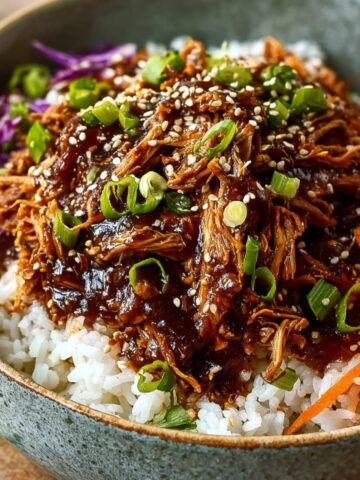 Slow Cooker Asian Pulled Pork served over white rice with a glossy dark sauce, garnished with sesame seeds and sliced green onions, accompanied by shredded purple cabbage and julienned carrots on the side