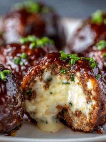 Cheese-stuffed meatballs with glossy barbecue glaze and chopped green onions on a white plate, with one meatball cut open revealing creamy melted cheese filling oozing out.