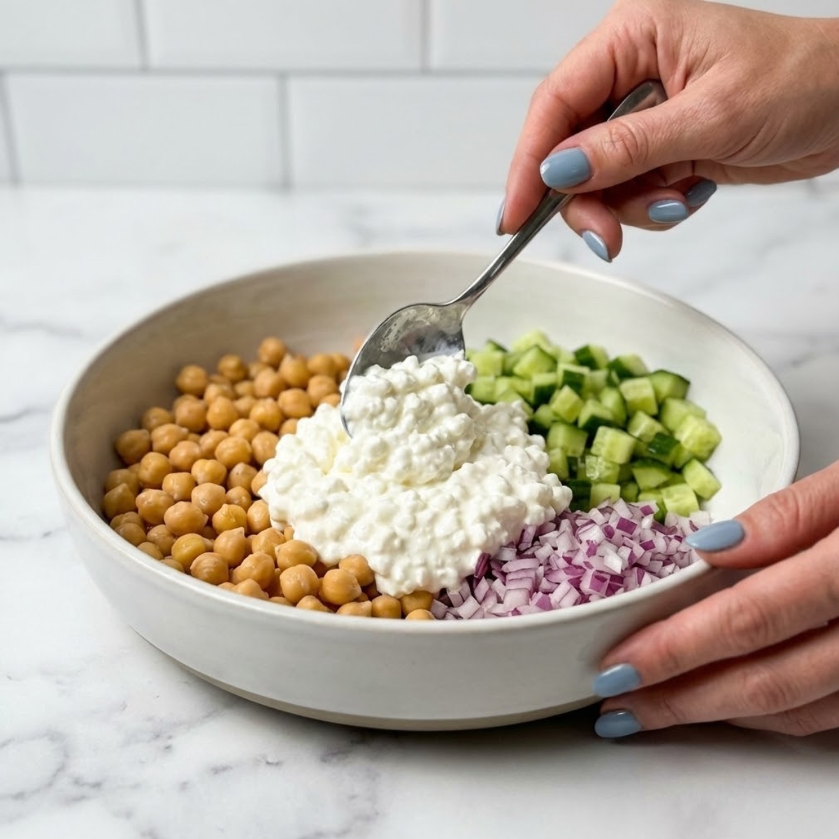 Close-up of hands arranging cottage cheese, chickpeas, cucumber, and onion in sections within a serving bowl.