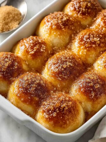 A white rectangular baking dish filled with golden-brown caramel apple pie bombs topped with coarse brown sugar crust, sitting on a marble surface with a spoon of brown sugar and a linen napkin alongside.