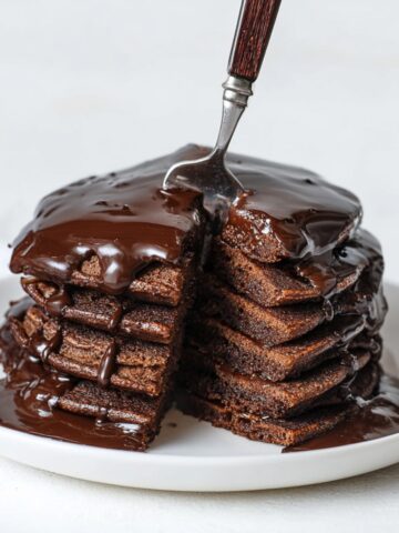Tall stack of double chocolate pancakes on a white plate with a fork cutting into the top, rich dark chocolate ganache sauce being poured over and dripping down the sides, pooling on the plate