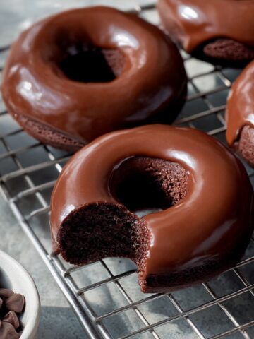 Chocolate protein donuts coated in a smooth, shiny chocolate glaze cooling on a metal wire rack, with one donut showing a bite taken out and a small bowl of chocolate chips nearby.