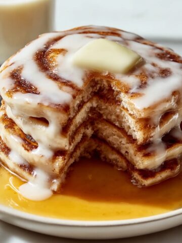 Stack of cinnamon roll pancakes with a wedge cut out on white plate, showing cinnamon swirl pattern throughout fluffy buttermilk pancakes, topped with melting butter pat and drizzled with cream cheese glaze, sitting in pool of maple syrup, with coffee cup blurred in background
