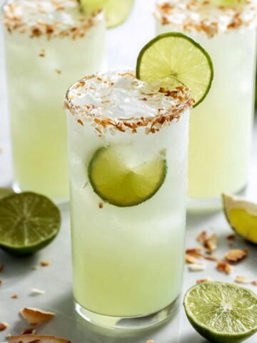Close-up of coconut lime refresher in a tall glass with toasted coconut flakes rimming the edge, pale green drink filled with ice and fresh lime wheel garnish, surrounded by halved limes and coconut flakes on white marble