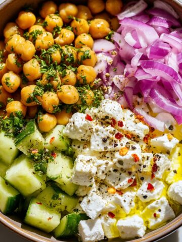 Mediterranean-style cottage cheese chickpea salad in a ceramic bowl with distinct sections: herb-coated chickpeas, diced cucumbers, cubed cottage cheese drizzled with olive oil and seasonings, and thinly sliced purple onions