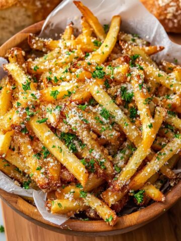 A wooden bowl lined with parchment paper filled with crispy air fryer french fries topped with grated parmesan cheese and chopped fresh parsley, with rustic bread loaves blurred in the background.