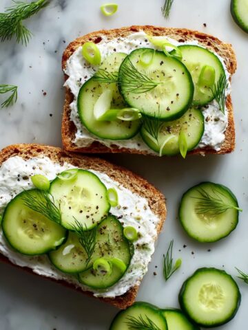 Two open-faced cucumber sandwiches on toasted whole wheat bread spread with herbed cream cheese, topped with thin cucumber slices, fresh dill, sliced green onions, and black pepper on a white marble surface