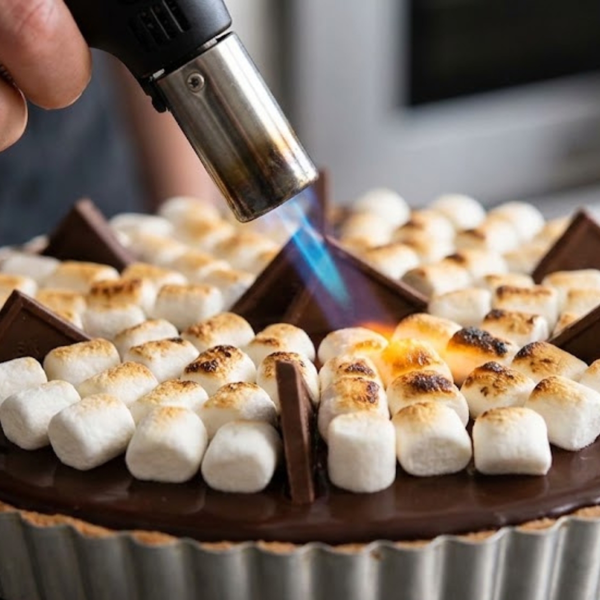 A square action shot of a kitchen torch browning mini marshmallows on top of a chocolate ganache S'mores Pie.