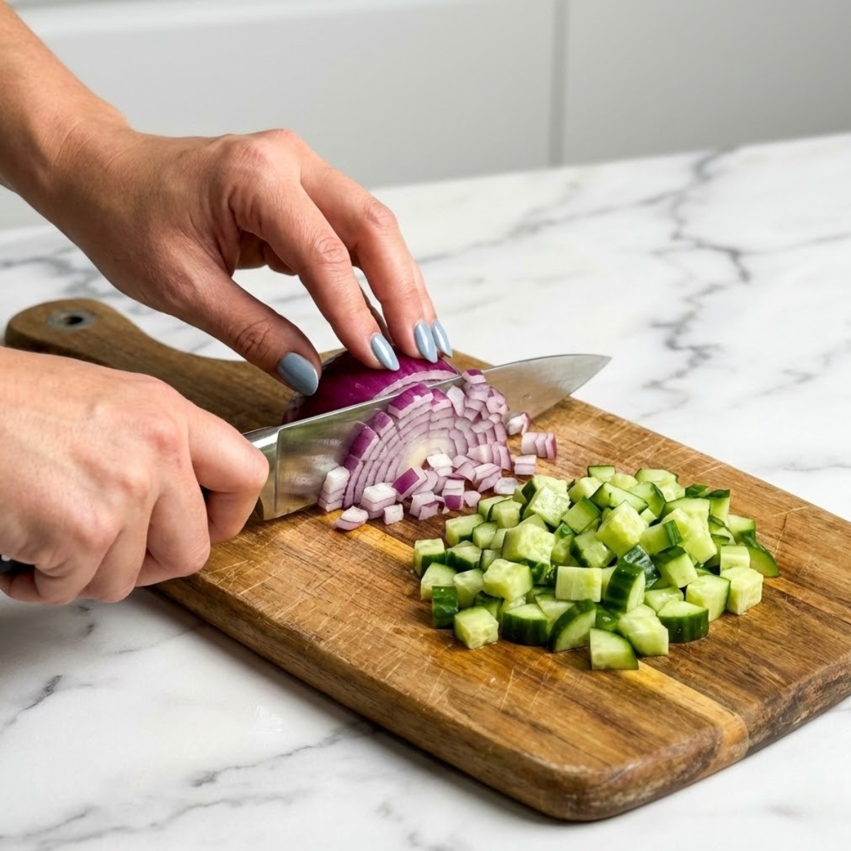 Extreme close-up of hands with fog blue nails dicing a red onion on a wooden cutting board.