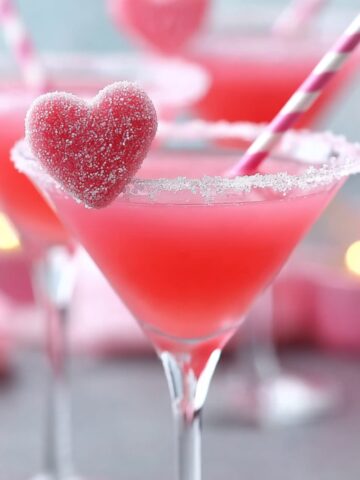 Valentine's Day Pink Sweetie Valen-Tini cocktail in a martini glass, filled with a bright coral-pink drink and rimmed with sparkling white sugar crystals. A red heart-shaped gummy candy covered in white sugar is perched on the rim, and a pink-and-white striped paper straw extends from the drink. The background is dreamily soft-focused, showing additional pink martinis, pink decorative elements, and warm golden candlelight bokeh, creating an ethereal Valentine's atmosphere.