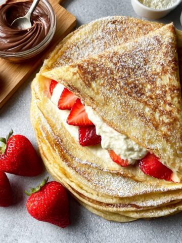 Stack of thin French crepes dusted with powdered sugar, folded to reveal filling of whipped cream and fresh sliced strawberries, with whole strawberries, chocolate spread in a glass bowl, and white bowls in the background