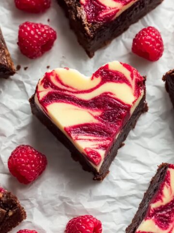 Heart Shaped Brownies arranged on white parchment paper with fresh raspberries, featuring fudgy chocolate brownie base with swirled cream cheese and raspberry topping in a marbled pattern, cut into heart shapes perfect for Valentine's Day