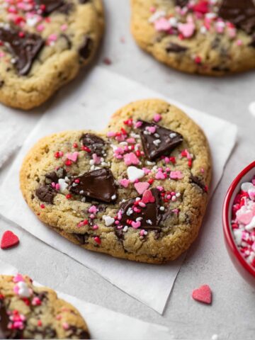 Overhead styled photo of heart-shaped chocolate chunk cookies on a light gray surface. The center cookie sits on a piece of parchment paper and is topped with melted dark chocolate chunks and a mix of pink, red, and white heart-shaped sprinkles. A red bowl filled with Valentine's sprinkles is positioned to the right, with scattered individual heart sprinkles around the surface. Additional cookies are visible in the background and corners of the frame, with a white linen napkin tucked beneath.