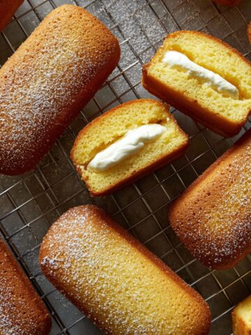Homemade Twinkies arranged on wire cooling rack, featuring golden oval-shaped sponge cakes dusted with powdered sugar, two cut in half to display piped white cream filling through the center of tender yellow cake