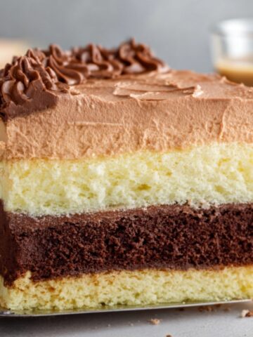 A close-up, side-profile shot of a square slice of Italian Love Cake being held on a metal cake server. The slice displays four distinct, horizontal layers: a thin vanilla sponge base, a thick dark chocolate cake layer, a thick pale-yellow ricotta cheese layer, and a top layer of light brown chocolate mousse. The top is finished with smooth frosting and a decorative piped border of dark chocolate buttercream swirls. The background is softly blurred, showing a hint of a beverage and neutral tones.