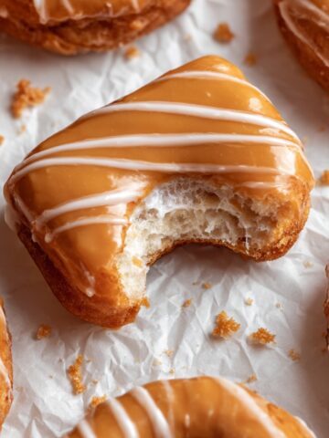 Maple donut bar with a bite taken out, showing fluffy interior texture, glazed with caramel-colored maple icing and drizzled with white icing stripes, arranged on white parchment paper with other donuts