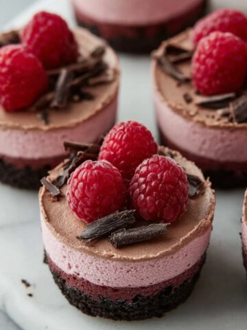 Close-up of no-bake raspberry chocolate mousse cups featuring dark Oreo cookie crust, ombre pink to chocolate layered mousse, topped with fresh raspberries and dark chocolate curls on white marble"