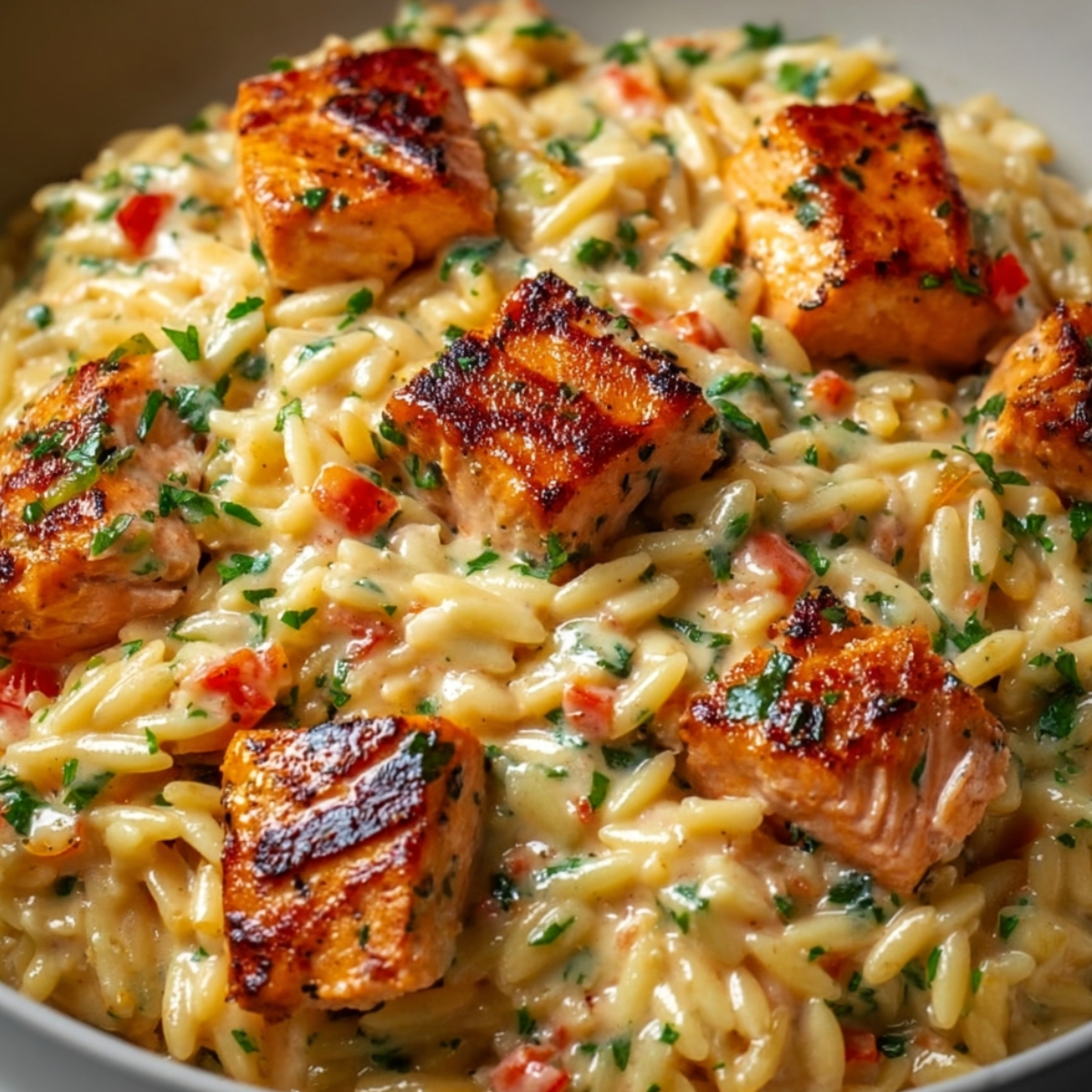 Close-up of Cajun chicken Alfredo orzo in a white bowl, featuring perfectly seared spice-crusted chicken pieces nestled in creamy orzo pasta studded with red bell peppers and fresh parsley