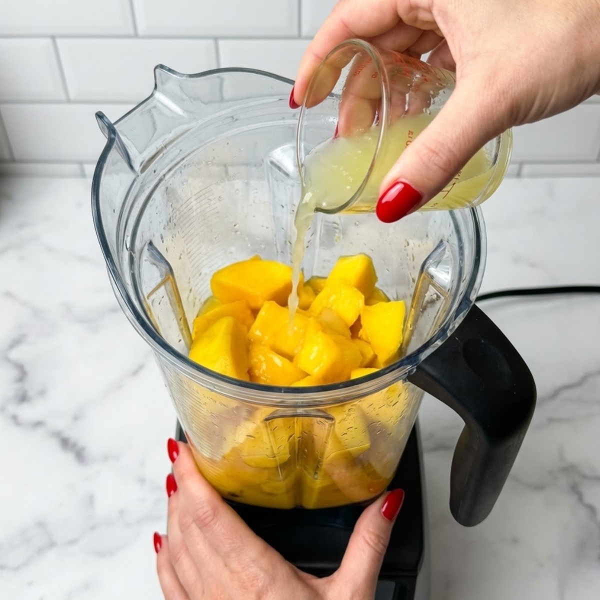 Extreme close-up of hands with red nails pouring lime juice over mango chunks in a blender.