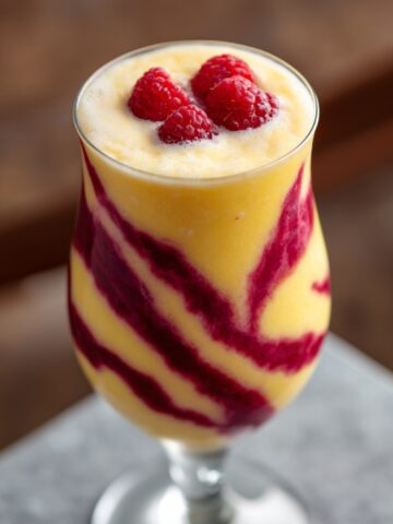 Raspberry Piña Colada in a stemless wine glass with a creamy yellow pineapple-coconut smoothie swirled with deep pink raspberry puree in decorative streaks, topped with three fresh raspberries, served on a white napkin