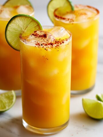 Three glasses of spicy mango limeade with bright golden-orange color, chili salt-rimmed glasses, filled with ice, garnished with fresh lime wheels, lime wedges scattered on white surface