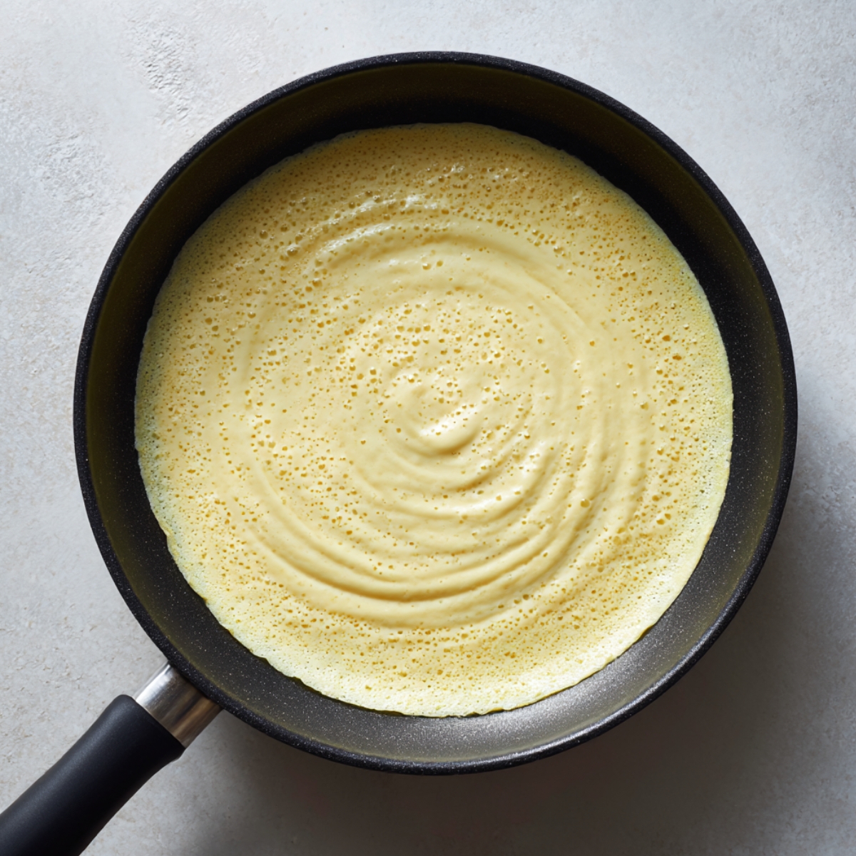 Overhead view of a hand swirling thin crepe batter in a nonstick pan to coat the bottom. Soft window light illuminates the cooking process for french crepes.