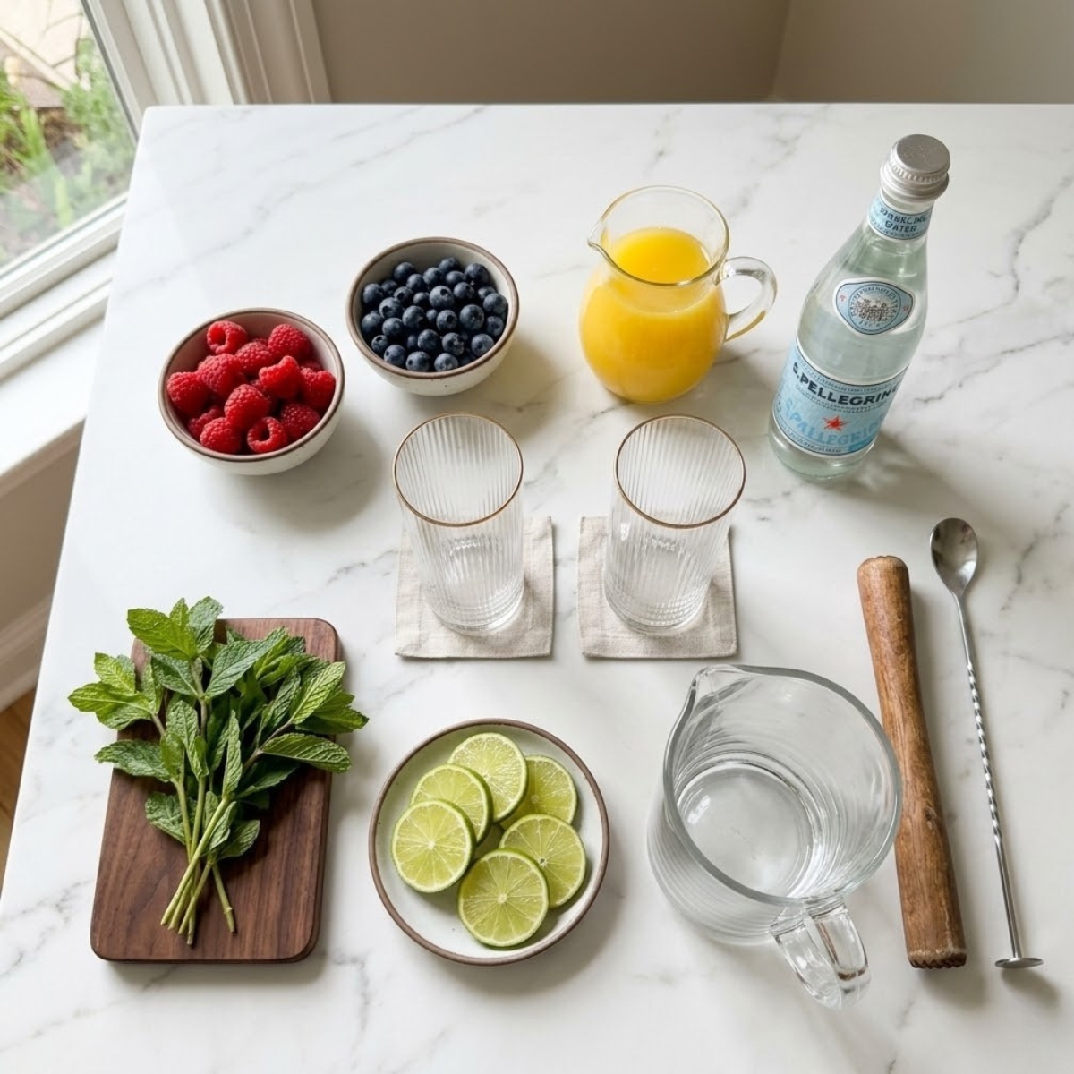 Overhead view of ingredients for a tropical mixed berry drink including raspberries, blueberries, pineapple juice, sparkling water, mint, and lime organized on a white marble counter without hands.