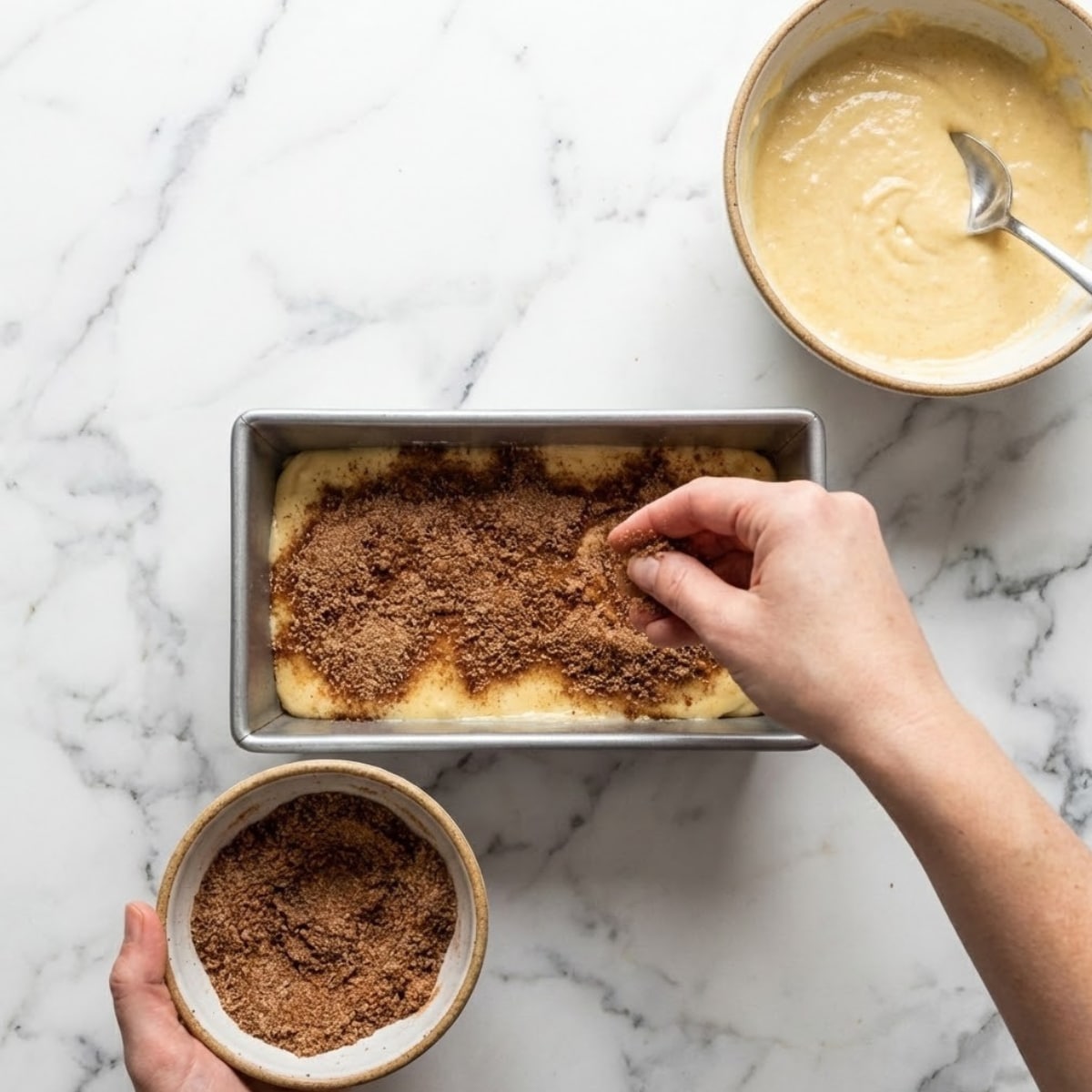 A top-down photograph showing the layering process for Amish Cinnamon Bread. A hand generously sprinkles a dark cinnamon-sugar mixture over a white marble kitchen surface. Bowls containing the remaining batter and cinnamon sugar sit beside the pan.