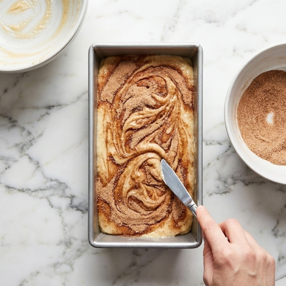 An overhead photograph showing a hand using a butter knife to gently swirl the cinnamon sugar topping into the batter of the Amish Cinnamon Bread just before baking. The metal loaf pan sits on a rustic wooden surface next to empty mixing bowls.