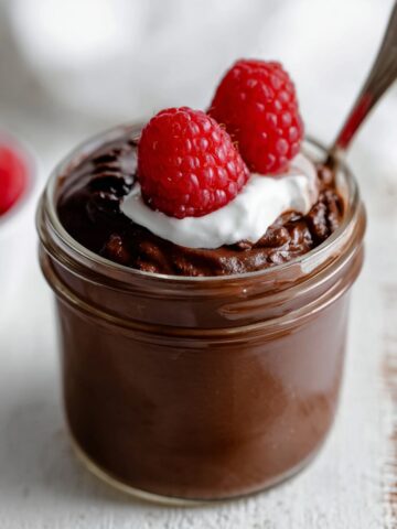A glass jar of smooth blended chocolate chia pudding topped with a dollop of whipped cream and two fresh raspberries, with a spoon resting in the jar and a bowl of raspberries blurred in the background.