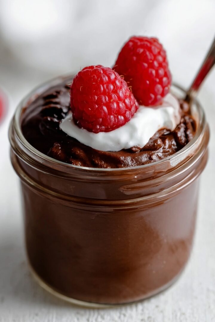 A glass jar of smooth blended chocolate chia pudding topped with a dollop of whipped cream and two fresh raspberries, with a spoon resting in the jar and a bowl of raspberries blurred in the background.