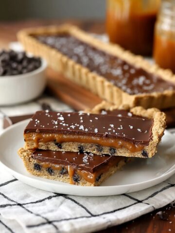 Chocolate Chip Salted Caramel Tart slices stacked on a white plate, revealing a chocolate chip cookie crust, gooey caramel layer, smooth chocolate ganache, and flaky sea salt.