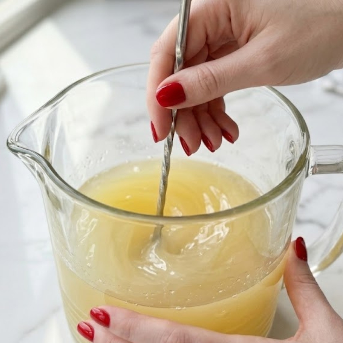 Close-up view of a young woman's hand with classic red nails using a stirring spoon to vigorously mix lemonade and pineapple juice in a glass pitcher on a white marble kitchen counter with natural daylight.