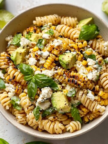 An overhead view of a speckled ceramic bowl filled with creamy street corn pasta salad — rotini pasta mixed with charred corn kernels, diced avocado, crumbled cotija cheese, and fresh basil leaves, with lime wedges and fresh basil scattered around the bowl on a white surface.