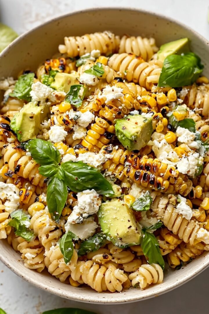 An overhead view of a speckled ceramic bowl filled with creamy street corn pasta salad — rotini pasta mixed with charred corn kernels, diced avocado, crumbled cotija cheese, and fresh basil leaves, with lime wedges and fresh basil scattered around the bowl on a white surface.