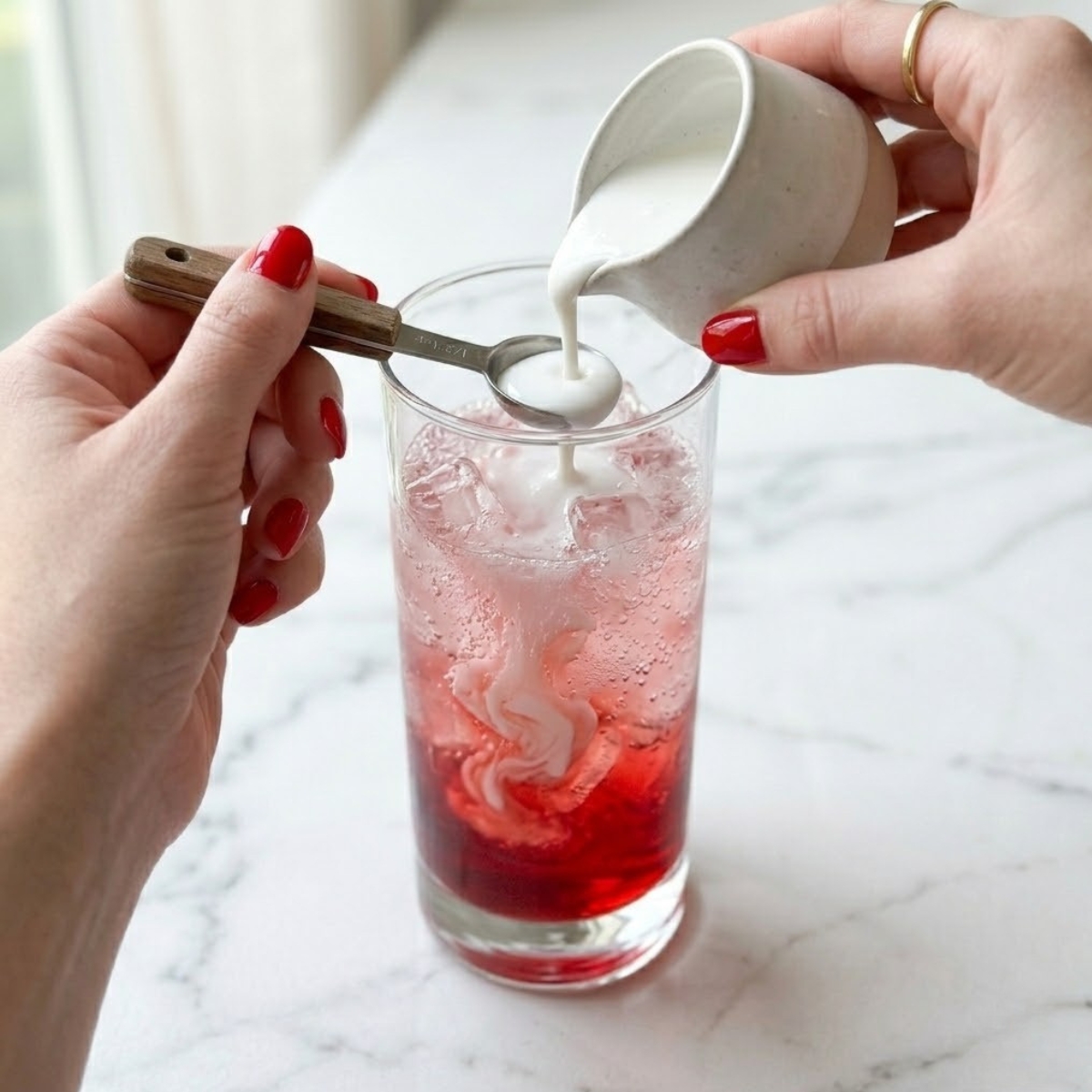 Close-up of a young woman's hands with polished classic red nails pouring thick white coconut cream over the back of a spoon into a tall glass of bubbly pink soda on a white marble kitchen counter, illustrating Step 3 of the recipe to create a dirty swirl.