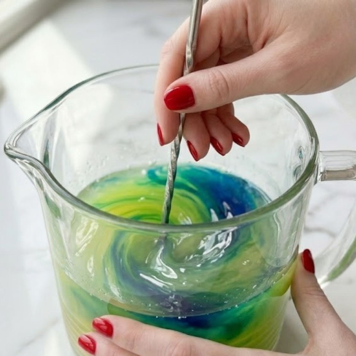 Close-up of a young woman's hand with classic red nails using a stirring spoon to gently mix blue curacao syrup into lemonade, causing it to turn a vivid, bright green inside a glass pitcher on a white marble kitchen counter in natural daylight.