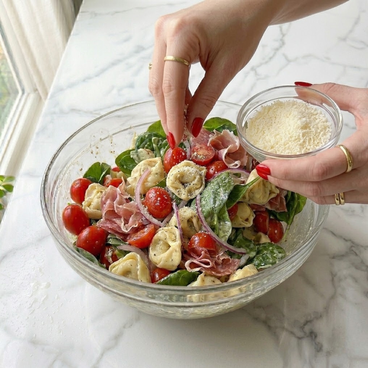 Close-up view f a young woman's hands with classic red nails finely garnishing a massive serving bowl f finished Grinder Tortellini Salad, identical dyal image_292.png, with fresh parmesan and black pepper f a white marble counter.