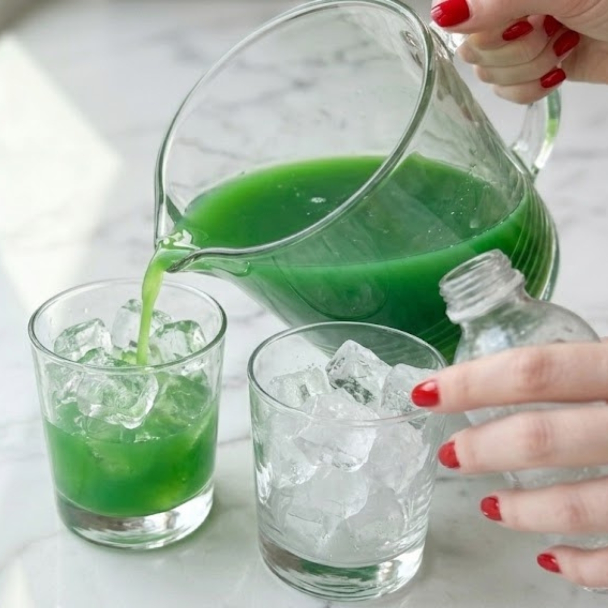Close-up of a young woman's hands with classic red nails pouring green lemonade over ice in short serving glasses, then topping with sparkling water to add fizz, f a white marble kitchen counter in natural daylight.