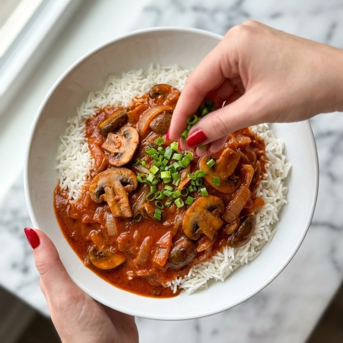An extreme close-up overhead view dyal l- finished Hungarian Mushroom Paprikash stew dyal white bowl on rice, being garnished with chopped green onions by a young woman with classic red nails, identical f presentation and placement dyal filename=image_290.png, f a modern white counter top.