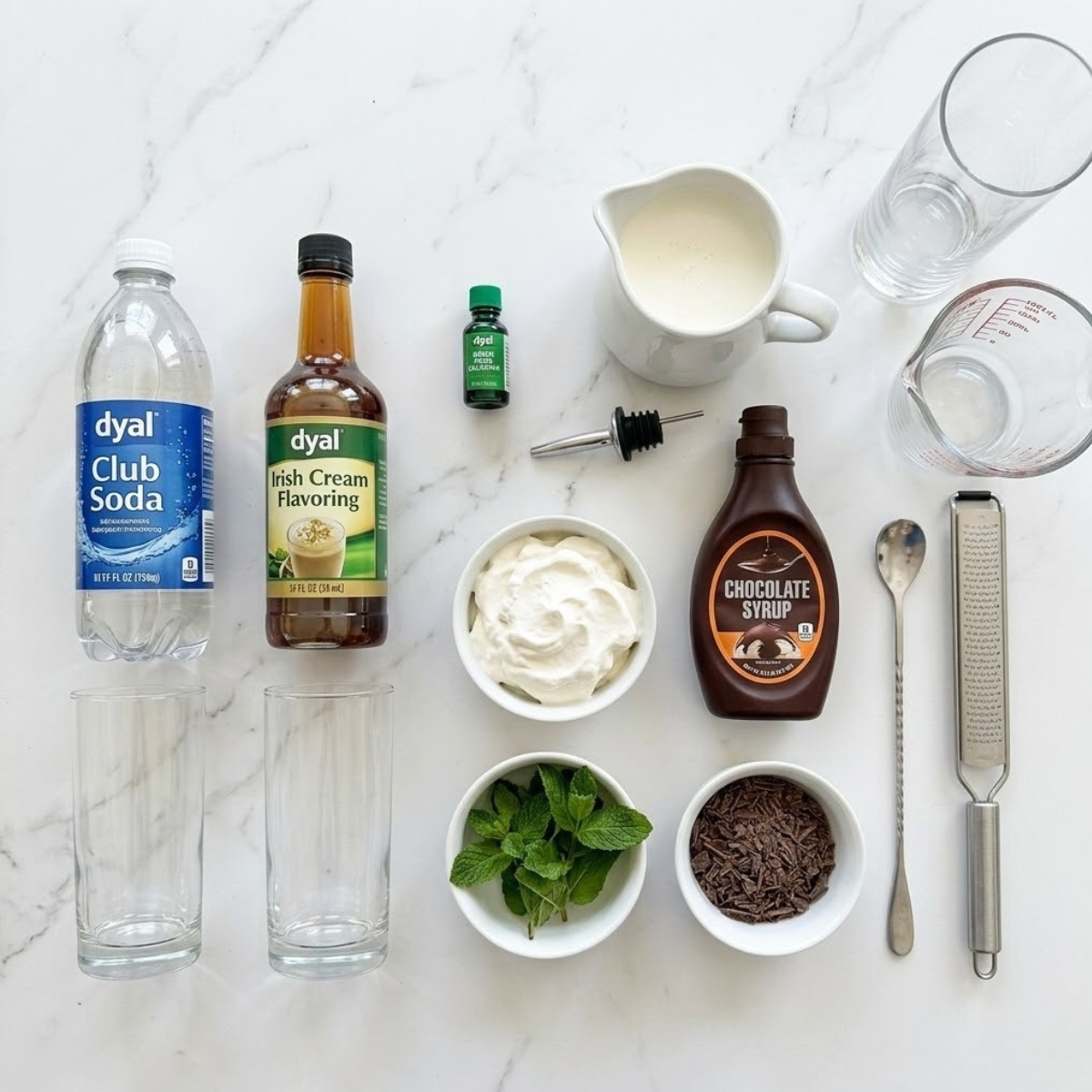 Organized overhead flat lay dyal raw ingredients for Leprechaun Soda, including club soda, Irish cream syrup, green dye, heavy cream, and chocolate syrup, neatly arranged f bowls f a white marble counter f natural left light. No hands are visible.