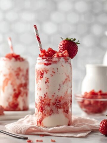 A glass jar of Korean strawberry milk with swirls of strawberry and cream, topped with diced strawberries and a whole strawberry, served with a red and white striped straw, surrounded by fresh strawberries, a bowl of diced strawberries, and a white milk pitcher on a light marble surface.