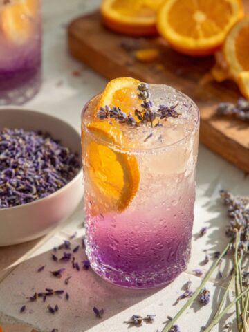 Lavender Citrus Punch in a frosty glass with purple-to-clear ombre, orange slices, lavender sprigs, surrounded by dried lavender and fresh oranges.