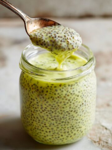 A glass jar of lemon curd chia pudding with a bright yellow-green hue and visible chia seeds, with a vintage spoon lifting a dripping spoonful above the jar, on a rustic stone surface.