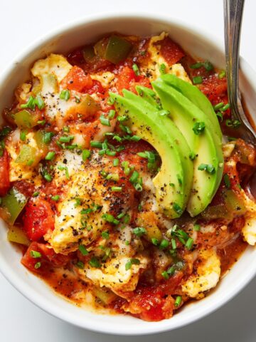 Overhead view of a white bowl of menemen Turkish egg scramble with eggs cooked in a chunky tomato and green pepper sauce, topped with sliced avocado, fresh chives, and cracked black pepper, with a spoon resting in the bowl.