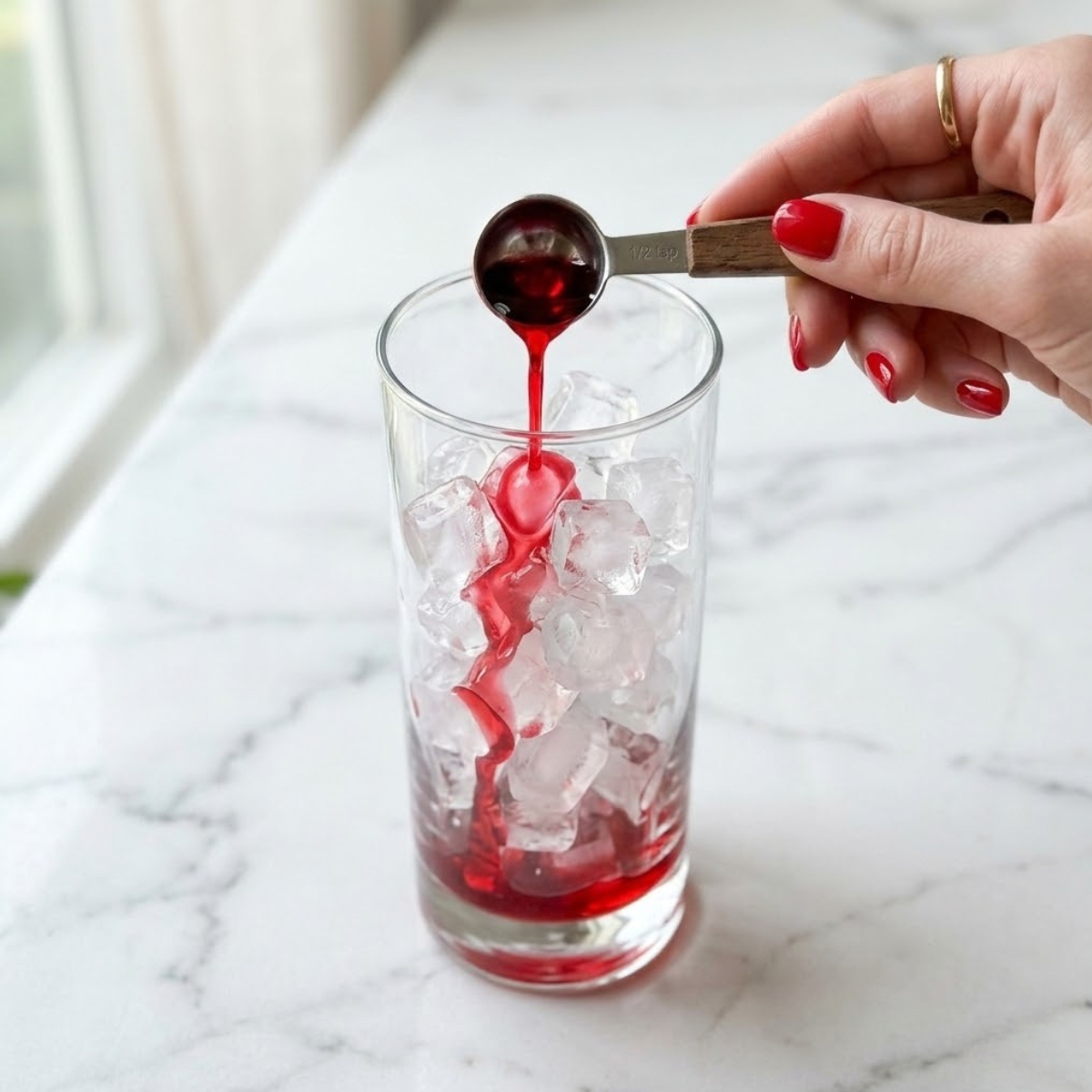 Close-up of a young woman's hand with polished classic red nails pouring bright red grenadine syrup from a spoon over ice into a tall serving glass on a white marble kitchen counter, demonstrating Step 1 of the recipe.