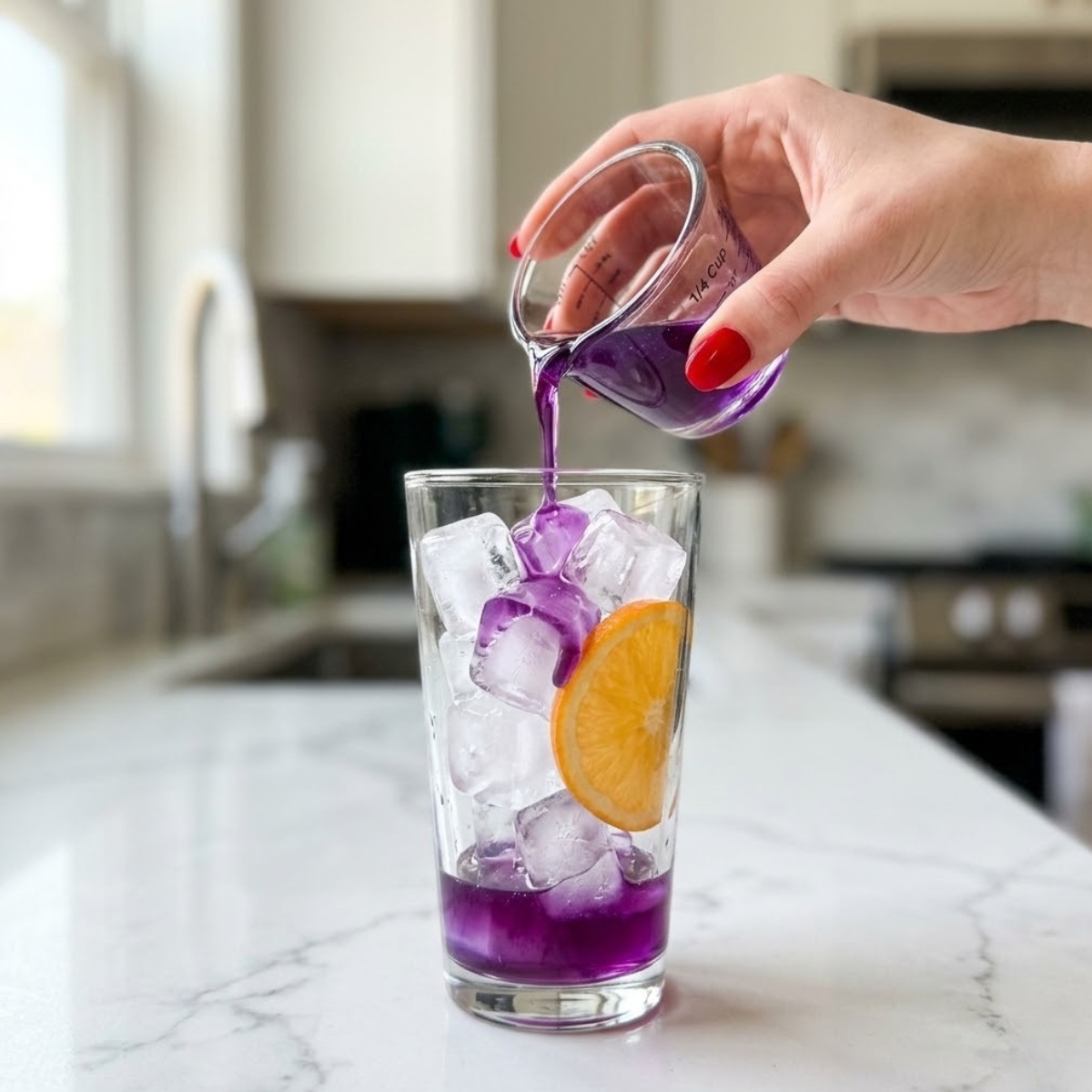 Close-up of a young woman's hand with red nails pouring vibrant purple lavender syrup over ice into a tall serving glass on a white marble kitchen counter.